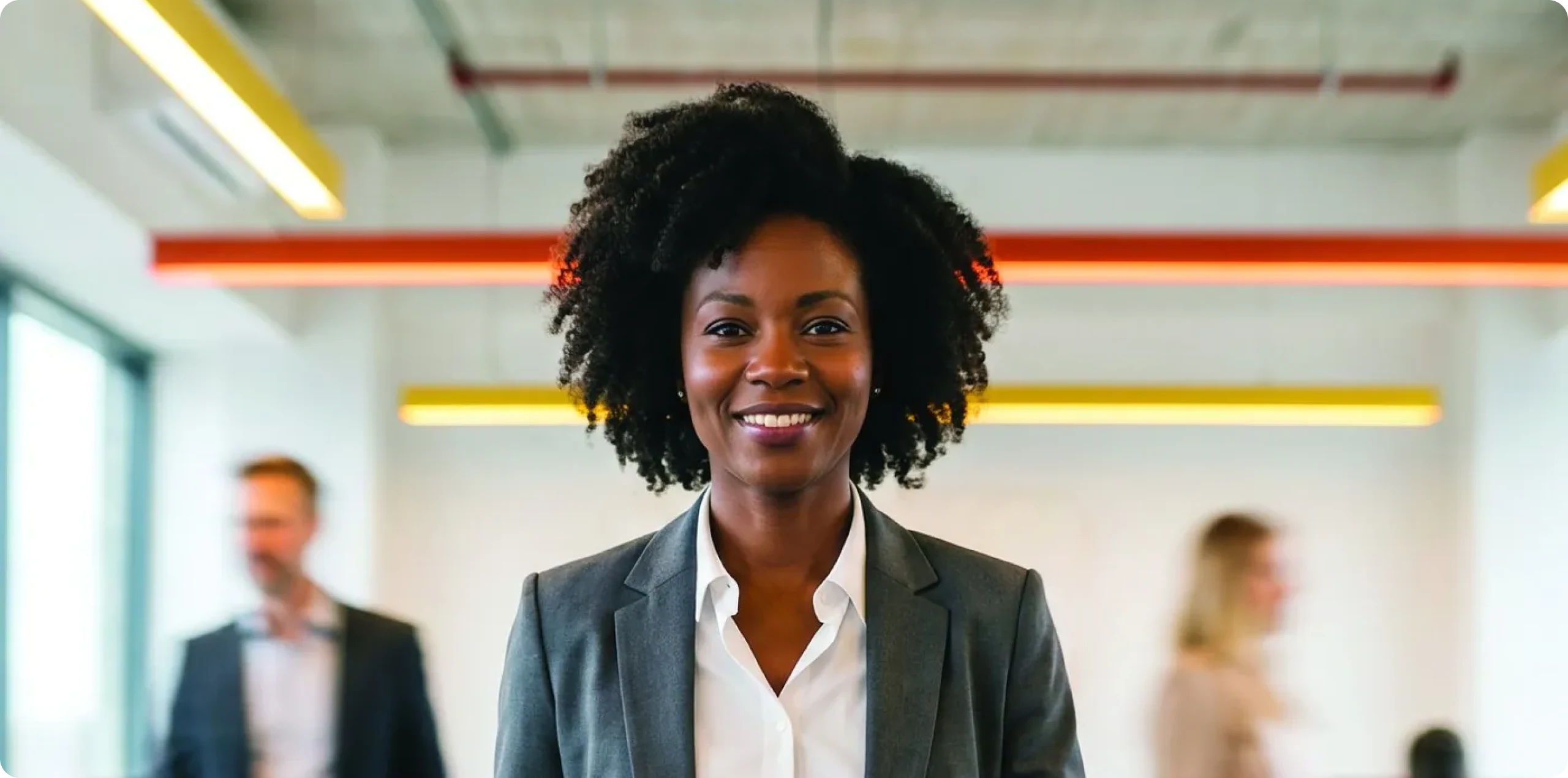 Executiva sorridente em ambiente corporativo, com colegas em movimento ao fundo.